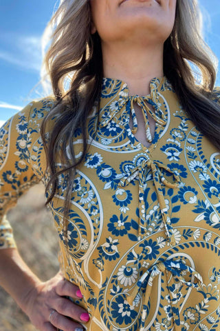 Sterling Kreek Women’s High Noon Honey Bow Top in mustard yellow paisley print with bow neckline and three-quarter sleeves.